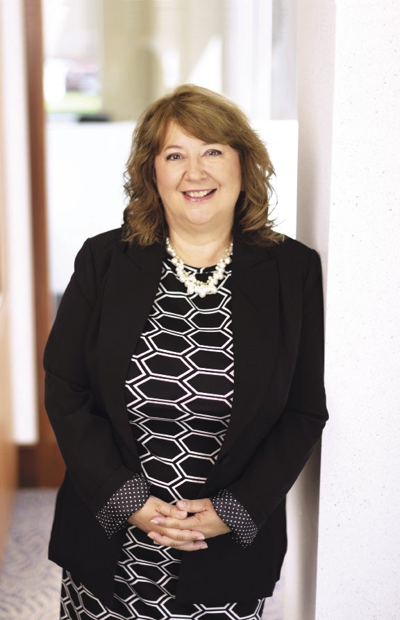 A woman wears a black blazer and black and white dress and leans against a white wall.