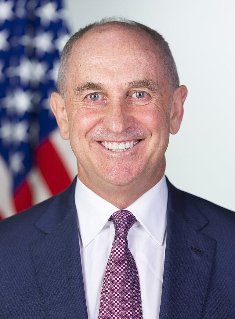 A man in a suit smiles in front of an American flag