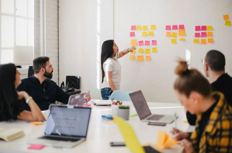 Woman in crowded board room points at sticky notes on wall