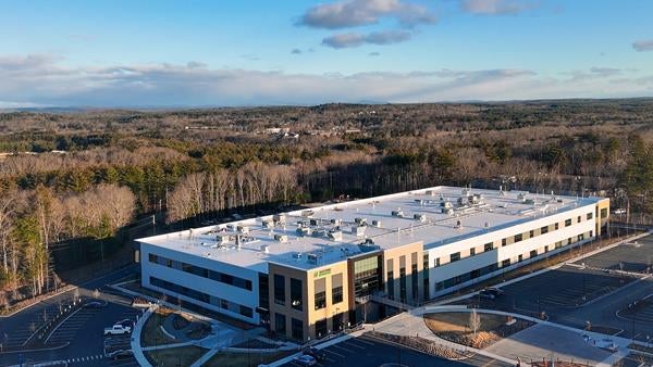 A large two-story industrial building sits in a parking lot in front of a forested area in an aerial photo. 
