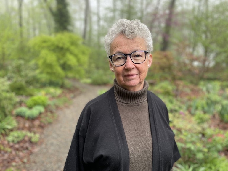 A woman with short grey hair wears glasses and a black cardigan while standing on a path in the woods.