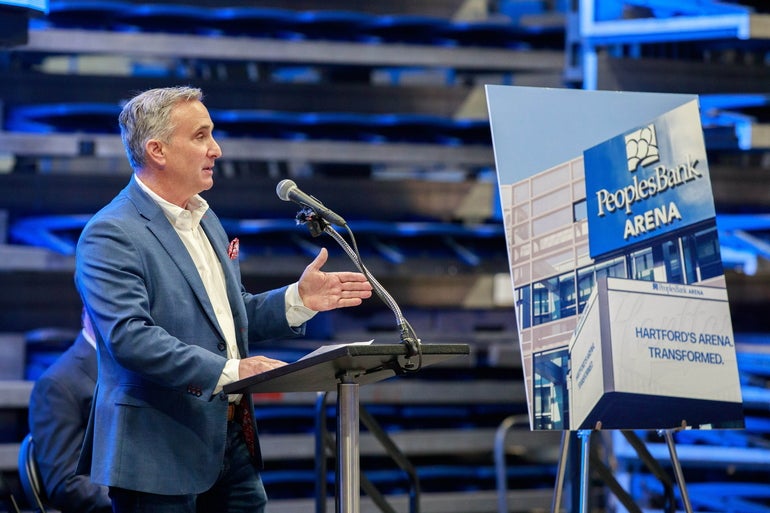 Man in suit stands at podium next to photo of PeoplesBank Arena in Hartford