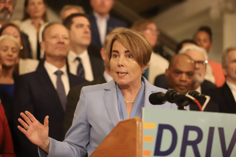 A woman wearing a powder blue suit stands talking at a podium with a group of people standing behind her.