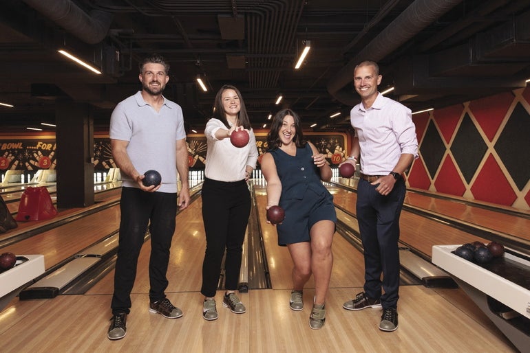 Four people standing on a candlpin bowling lane holding bowling balls