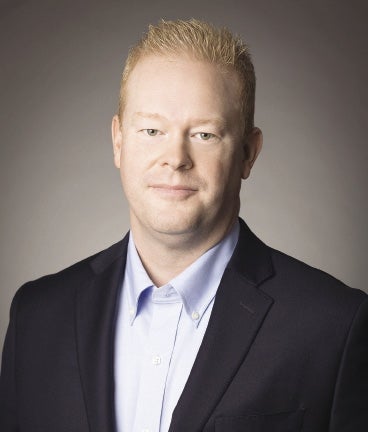 A man with short blonde hair wears a black suit jacket and light blue button up shirt.