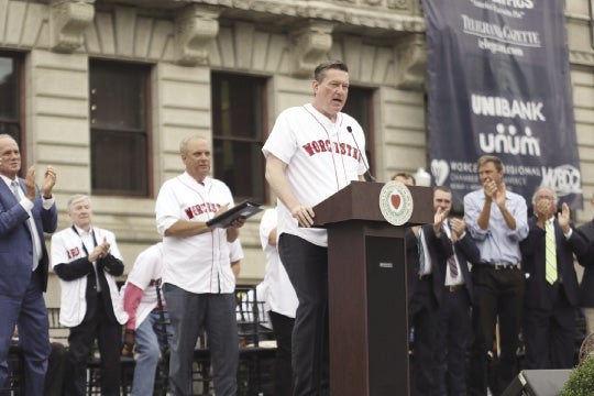 A man in a baseball jersey stands at a podium in front of a crowd on a stage.