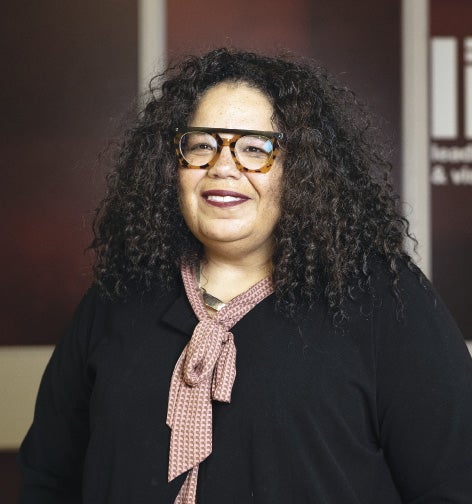A woman with dark curly hair wears a black top and black and tortoise glasses.
