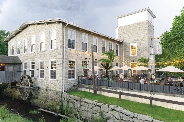 The historic mill made of stone, with a water wheel and covered bridge overlooks the brook that once powered it. Outside there is a patio with patrons dining under string lights at the GlenPharmer Distillery. 