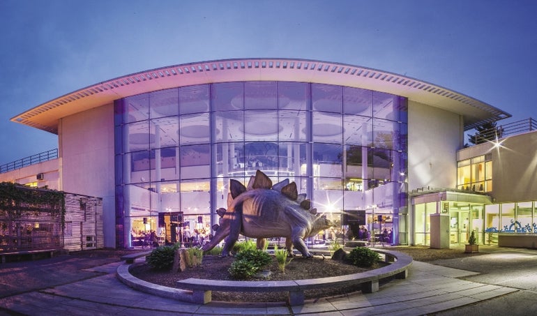 A building with a large glass front sits behind a statue of a dinosaur.  