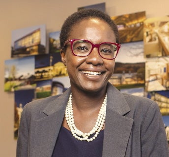 A woman with short dark hair wears a grey blazer, pearl necklace, and red glasses.