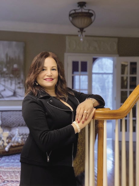 A women with brown hair wears a black jacket and skirt, leaning against a stair railing