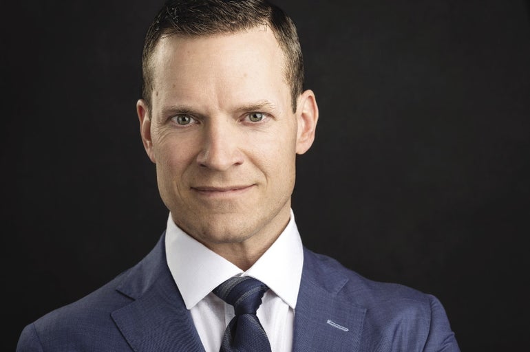 A man in a blue suit, white shirt, and blue tie stares intently at the camera.