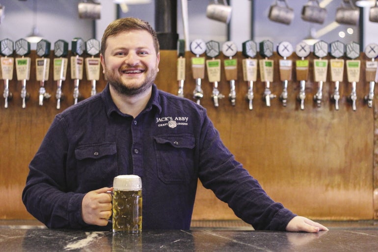 A man with a beard wearing a blue shirt holds a beer and stands in front of a row of taps.