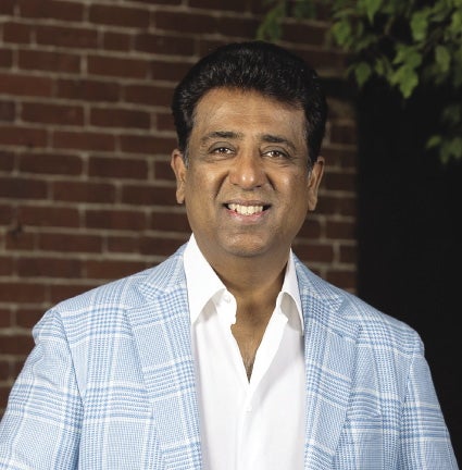 A man with short dark hair wears a light blue and white plaid suit jacket and a white button down.