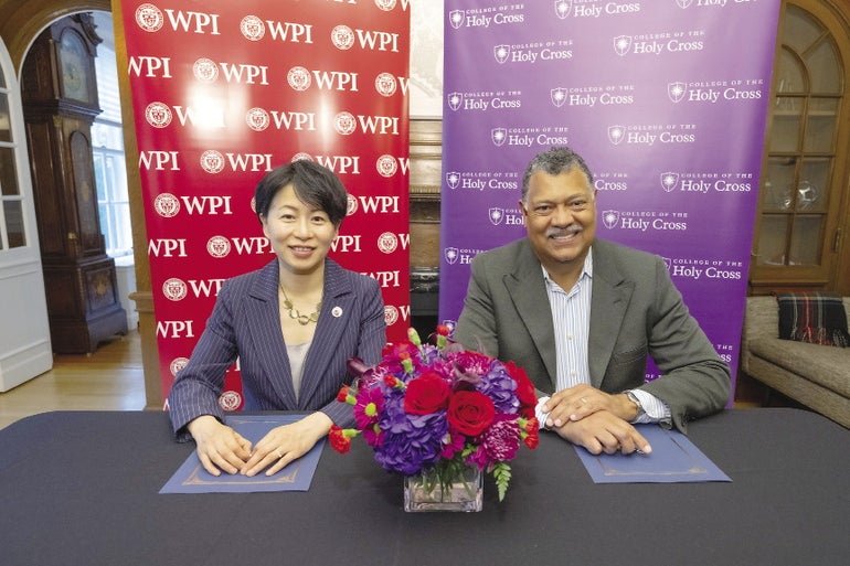 A woman and a man sit at a table in front of banners advertising WPI and College of the Holy Cross. 