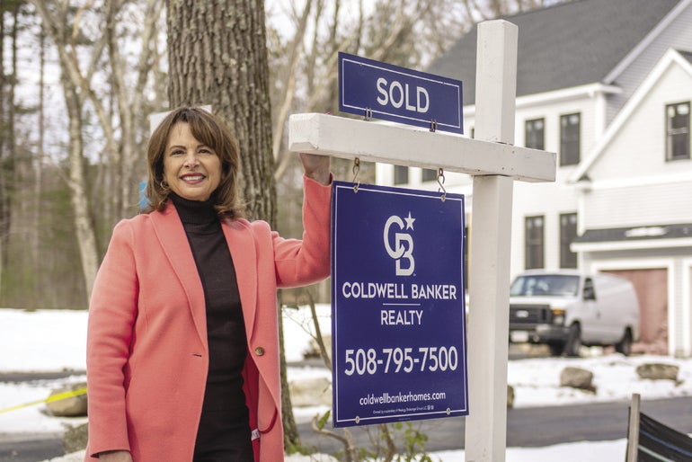 A woman stands in a pink blazer next to a real estate sign