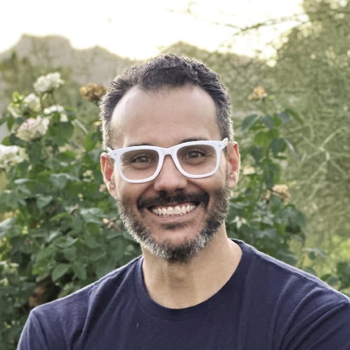 A man with dark hair and a beard wears white-rimmed glasses.