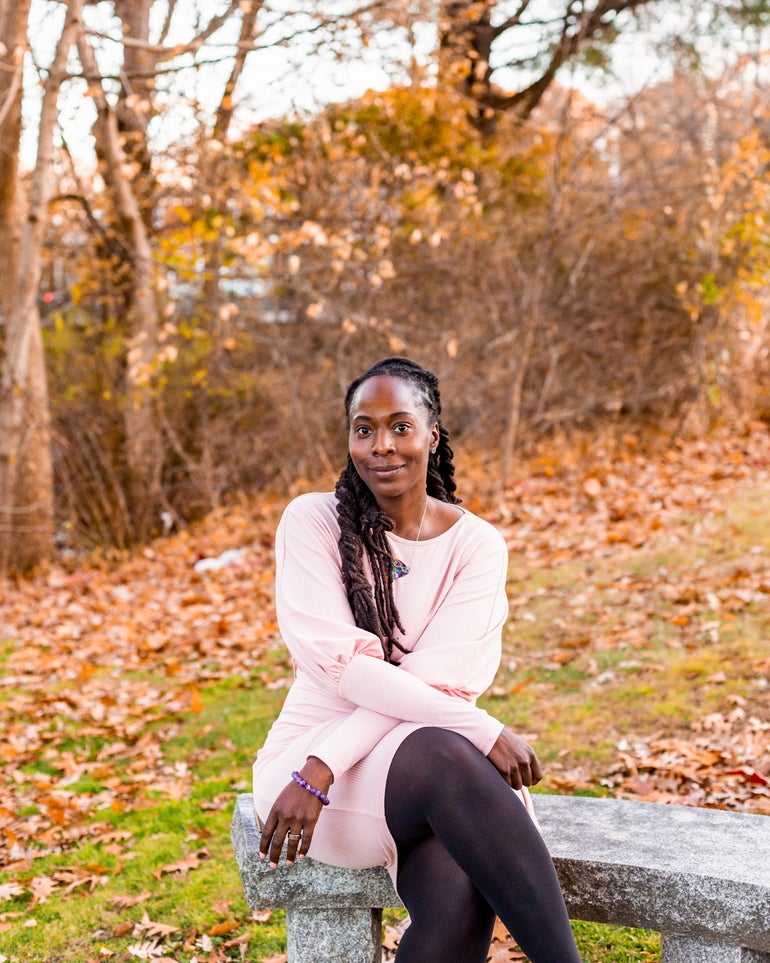 A woman with long dark brown hair wears a light pink dress and dark tights sitting on a grey stone bench in front of fall foliage.