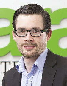A man with glasses wearing a suit sits in front of a green logo.