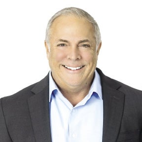 A man with gray hair wears a blue shirt and blue blazer