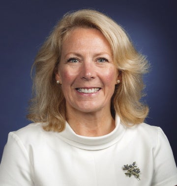 A woman with shoulder-length blonde hair wears a white blouse and a leaf broach.