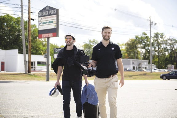 two men carrying equipment walk toward the camera in a parking lot