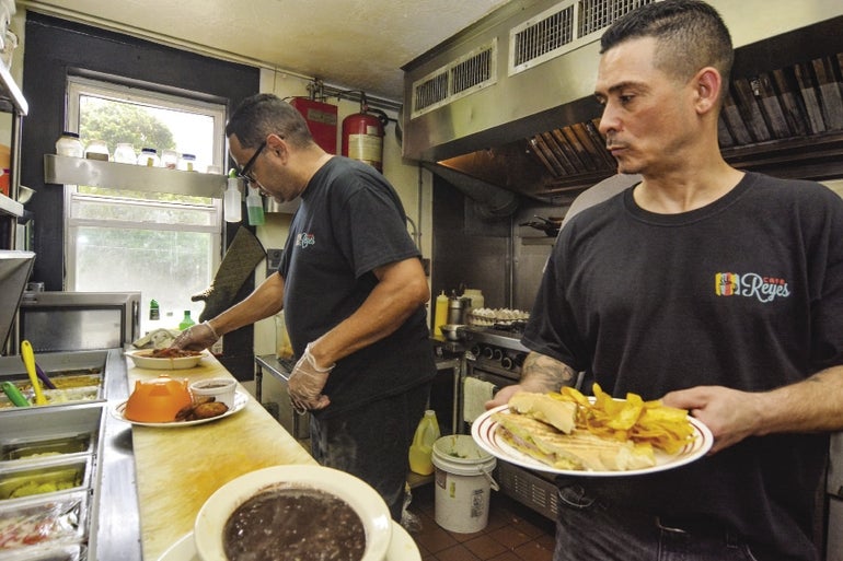 Two men in black shirts with the a small logo "Café Reyes" logo handle plates of food in the café kitchen  