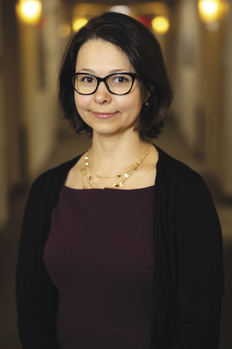 A woman wish short dark hair wears black glasses, a dark purple top, and a black cardigan.