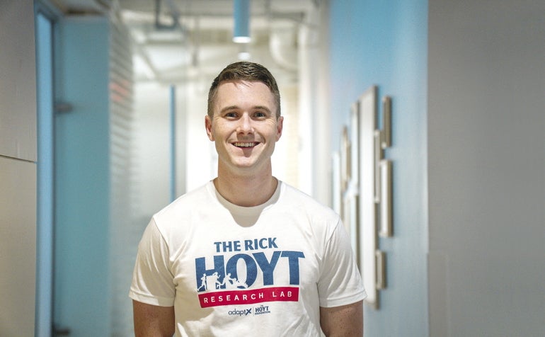 A young man wears a white T-Shirt that says "The Rick Hoyt Research Lab".