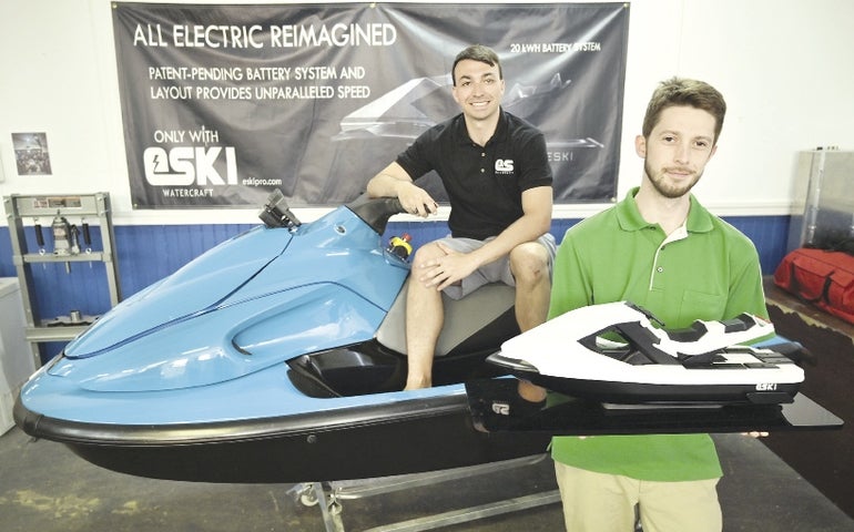 A man sits on a blue personal watercraft while another man hold a model of a futuristic looking personal watercraft