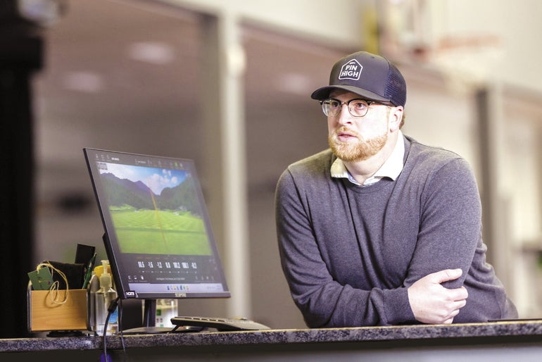 A man in a baseball cap leans on a counter with a computer screen next to him