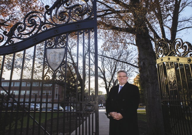 A man in a suit and black overcoat stands in front of an open cast-iron gate.
