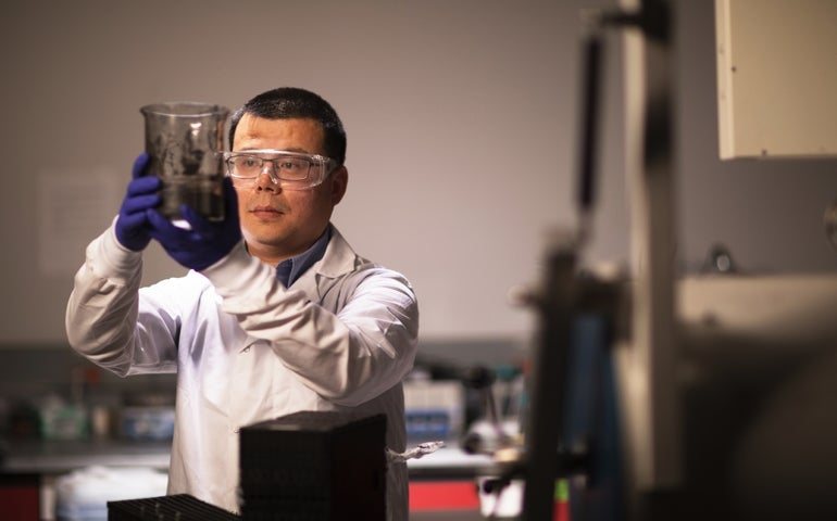 A man wearing a white lab coat and safety goggles holds up a beaker