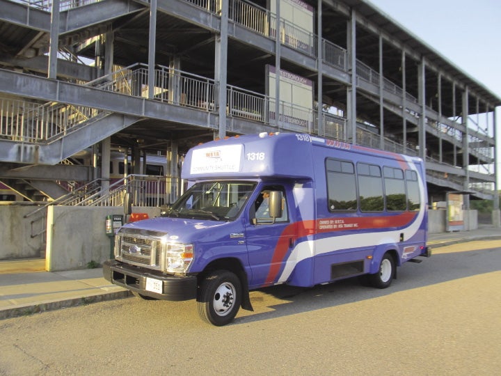 A small blue passenger bus with a large white and red stripe sits outside of a three-layer train station.