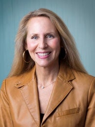 A woman with long blonde hair wears hoop earrings and a light brown leather jacket.