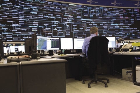 A man in a blue shirt sits at a computer in front of a large screen tracking energy flow.
