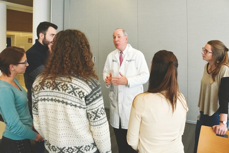 A man in a white lab coat speaks to five people in a semicircle around him.