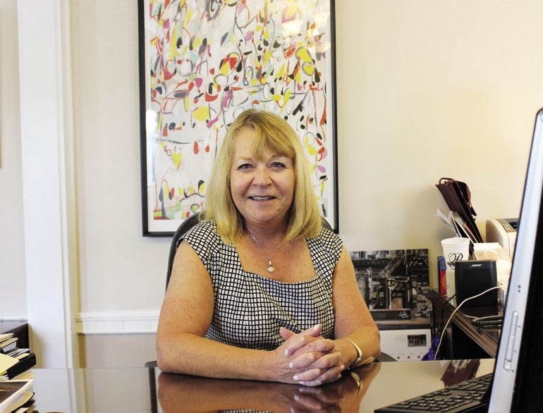 A woman with shoulder length blonde hair and bangs wears a black and white top sitting at a desk with her hands held together..