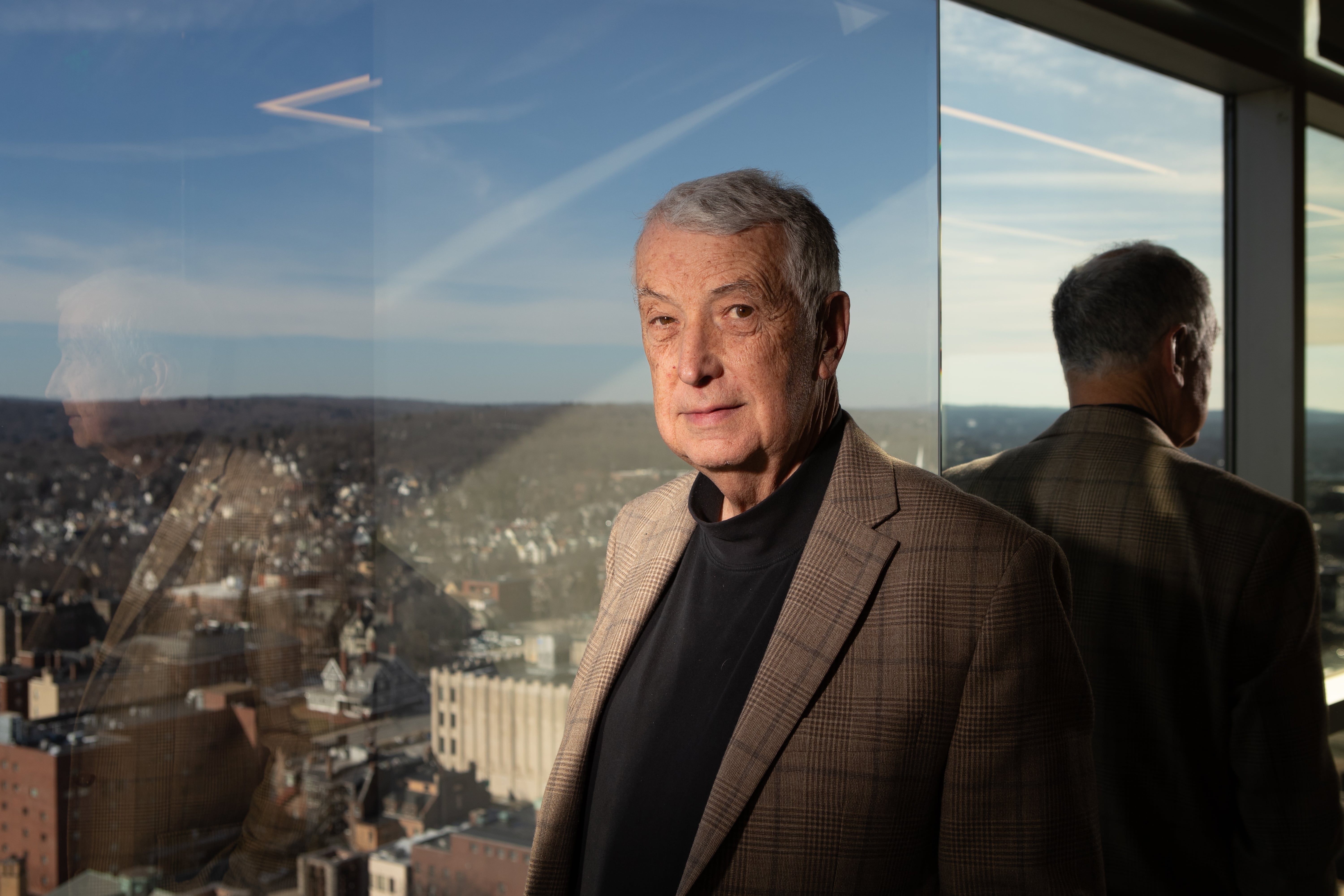 A man wearing a tan jacket and black turtleneck stares at the camera.