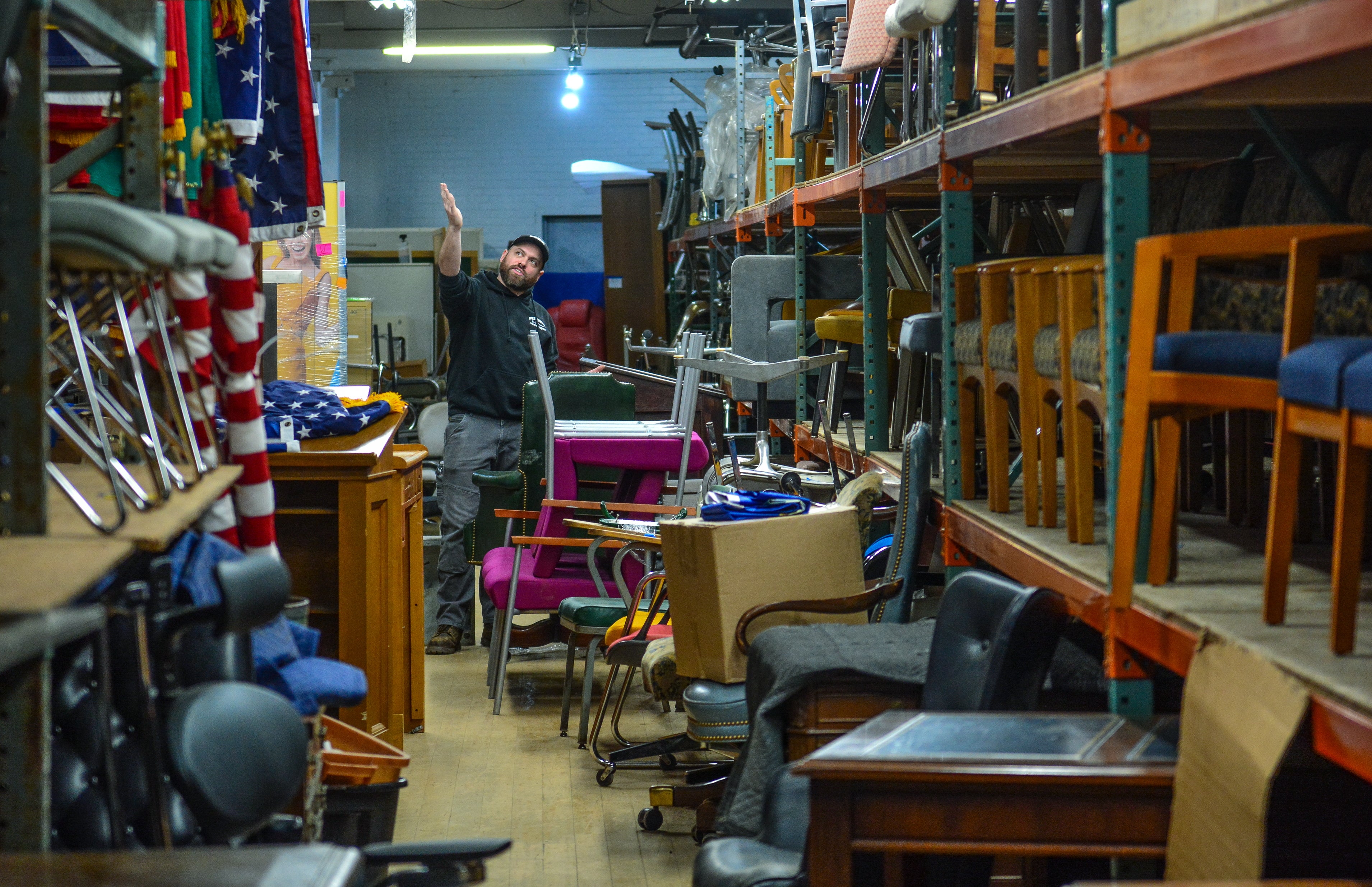 A man points at something above him as he stands among a large amount of office chairs and other furniture