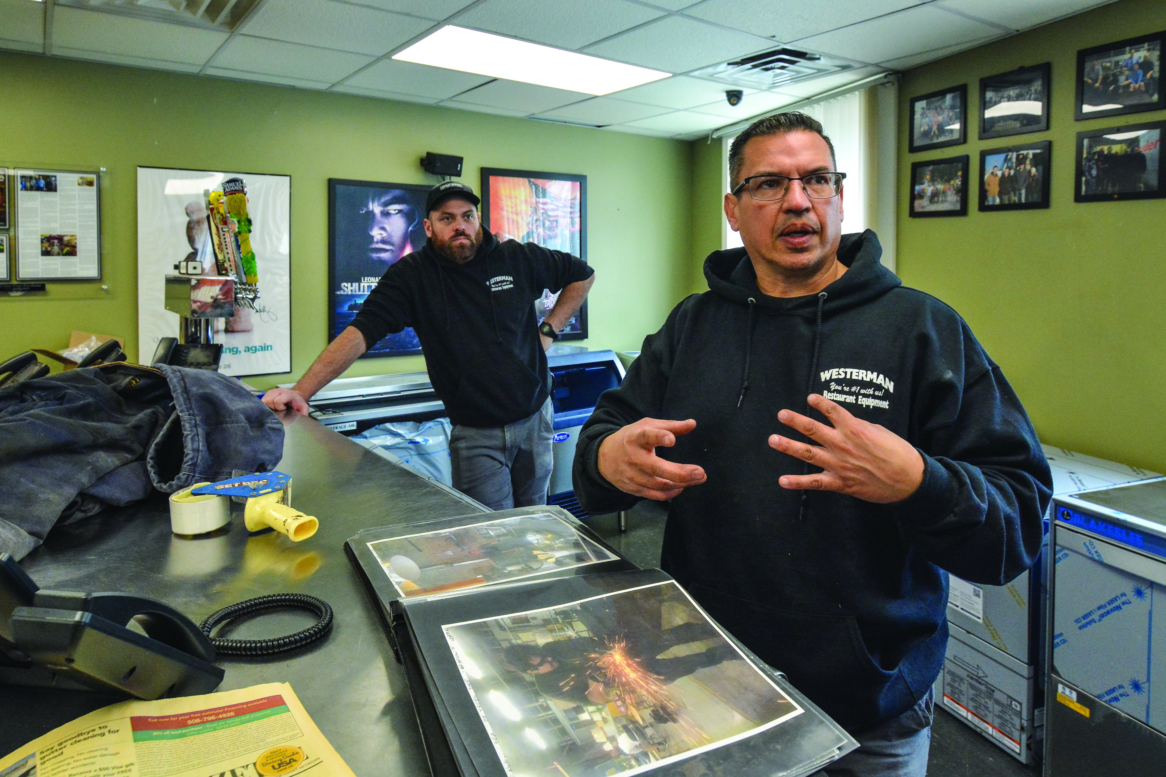 A man speaks to the camera while standing over a scrapbook, as another man looks on from behind