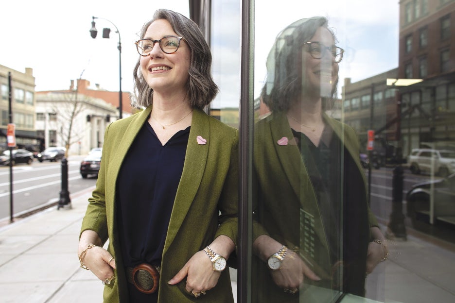 A woman in a green jacket and blck blouse leans against a window, smiling while looking off into the distance