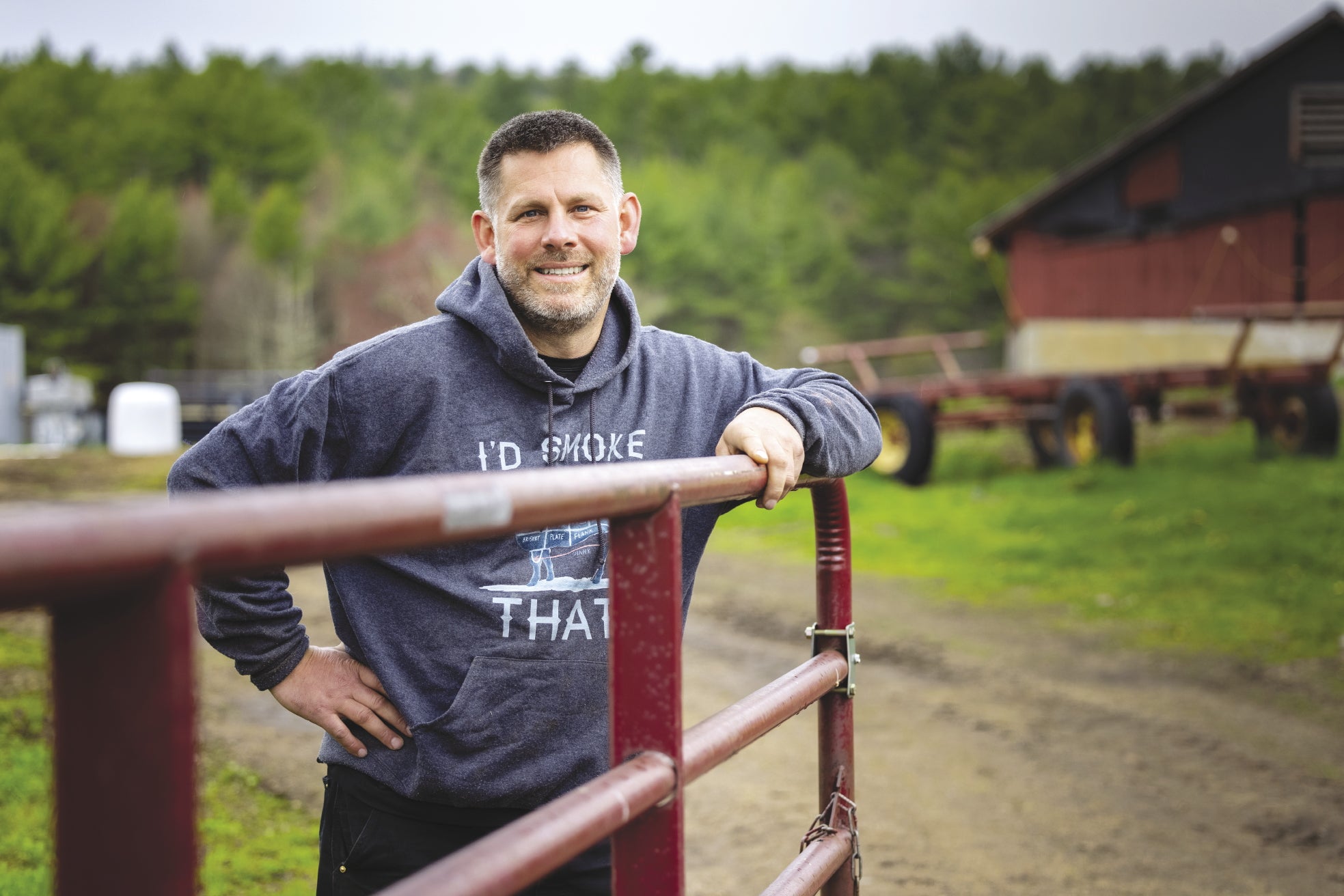 A man in a hooded sweatshirt smiles while leaning on a farm gate