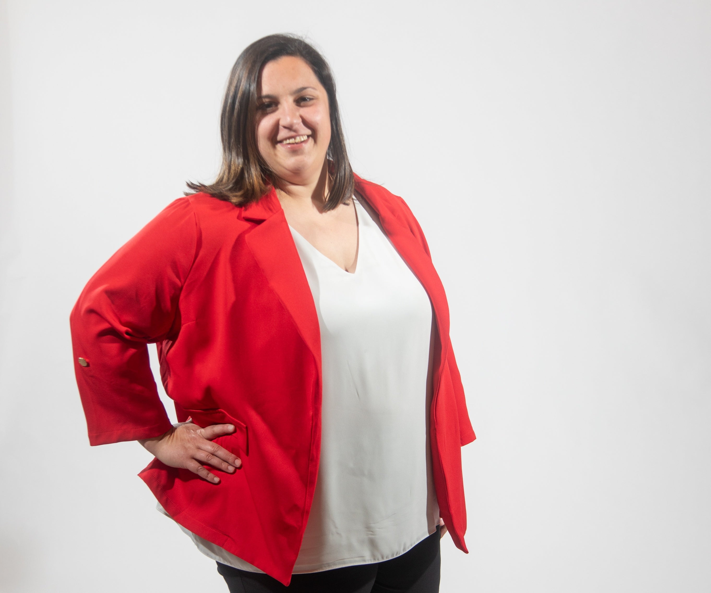 A woman with shoulder length brown hair wears a white top and red blazer with her hand on her hip.