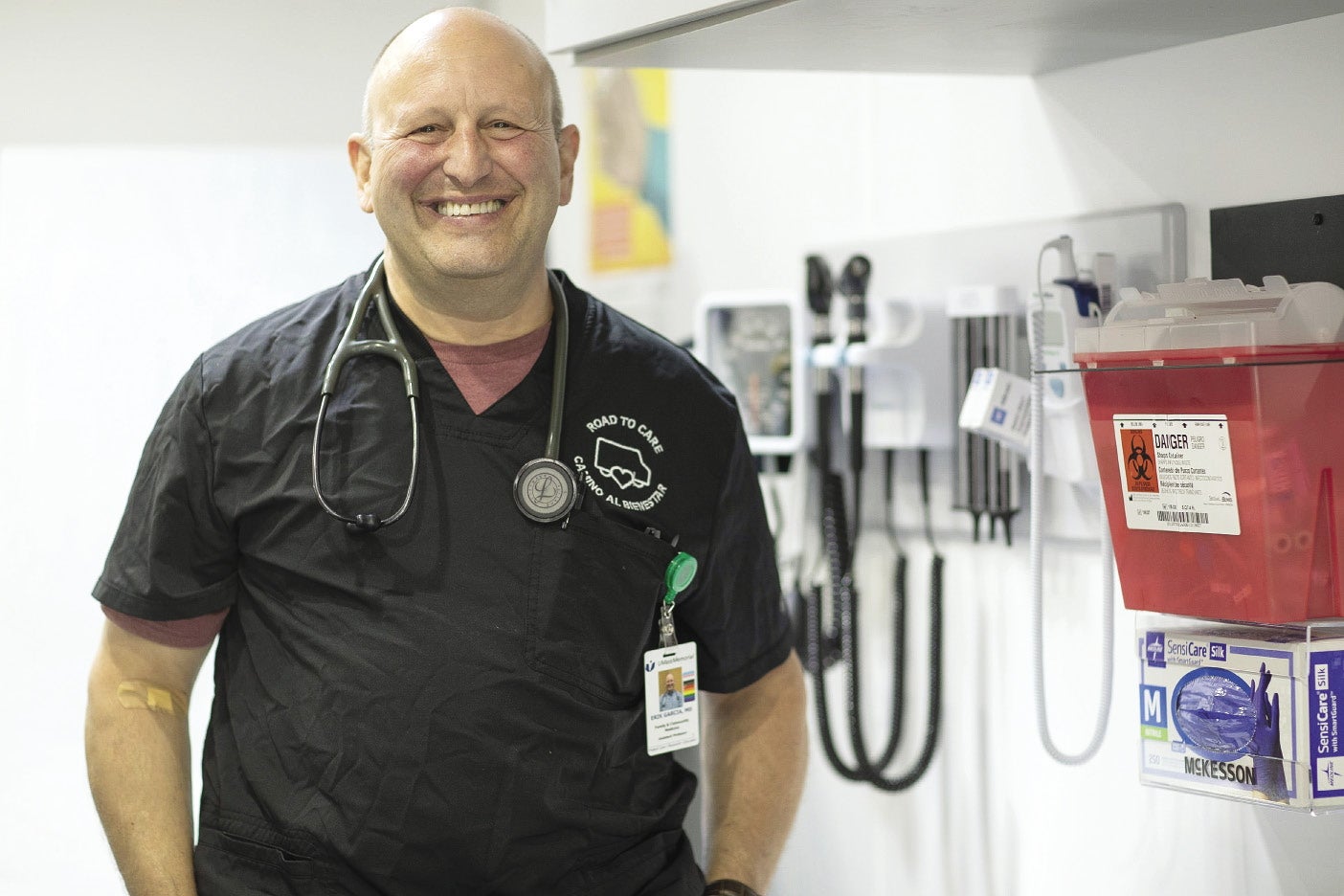 A bald man stands in a medical room with medical supplies on the wall while wearing black scrubs with a stethoscope around his neck.