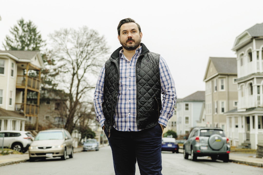 A man in a vest, plad shirt and jeans stands with his hands in his pocket in the middle of a residential street