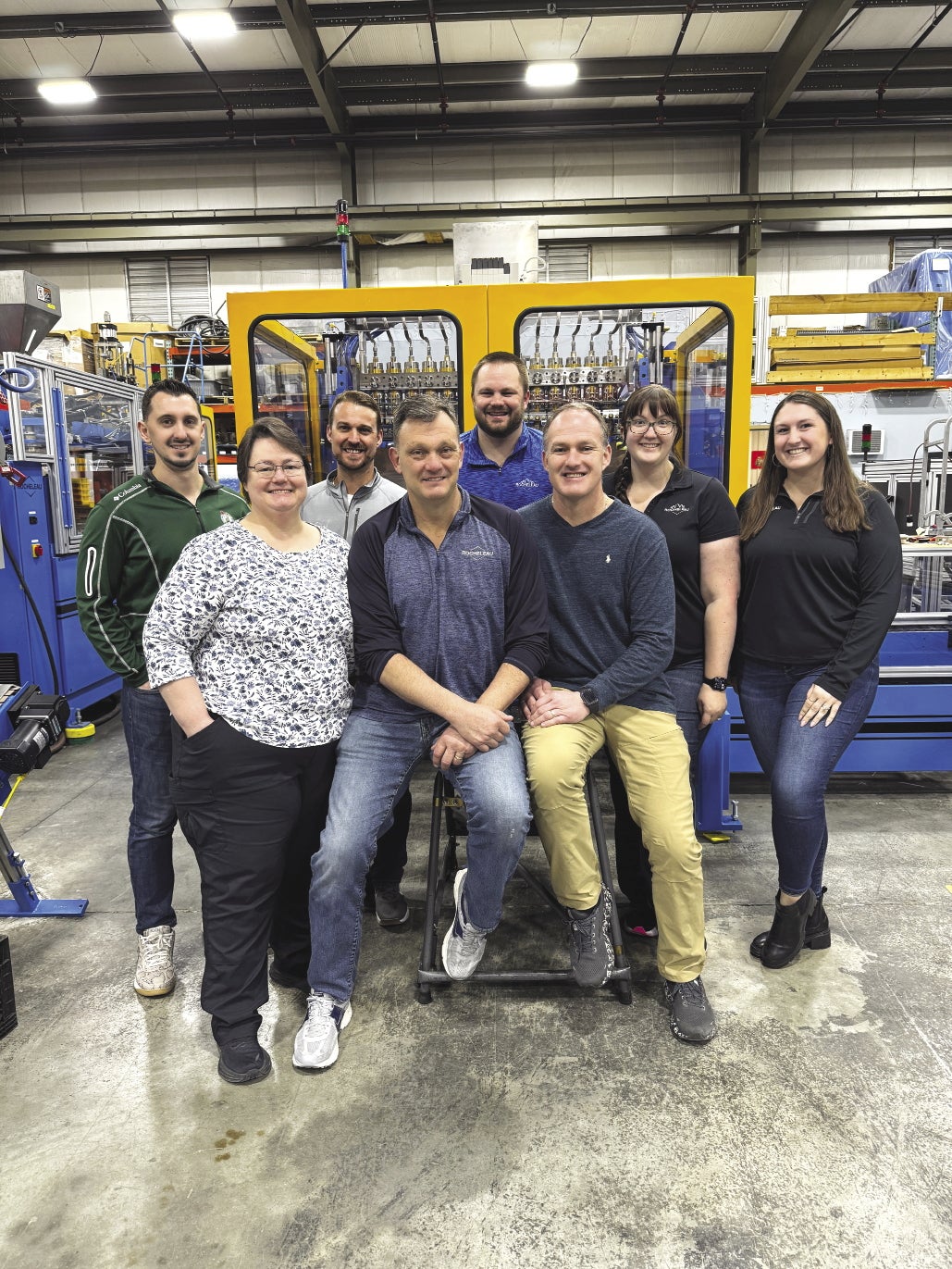 Eight people stand together in front of manufacturing machinery on a cement floor.