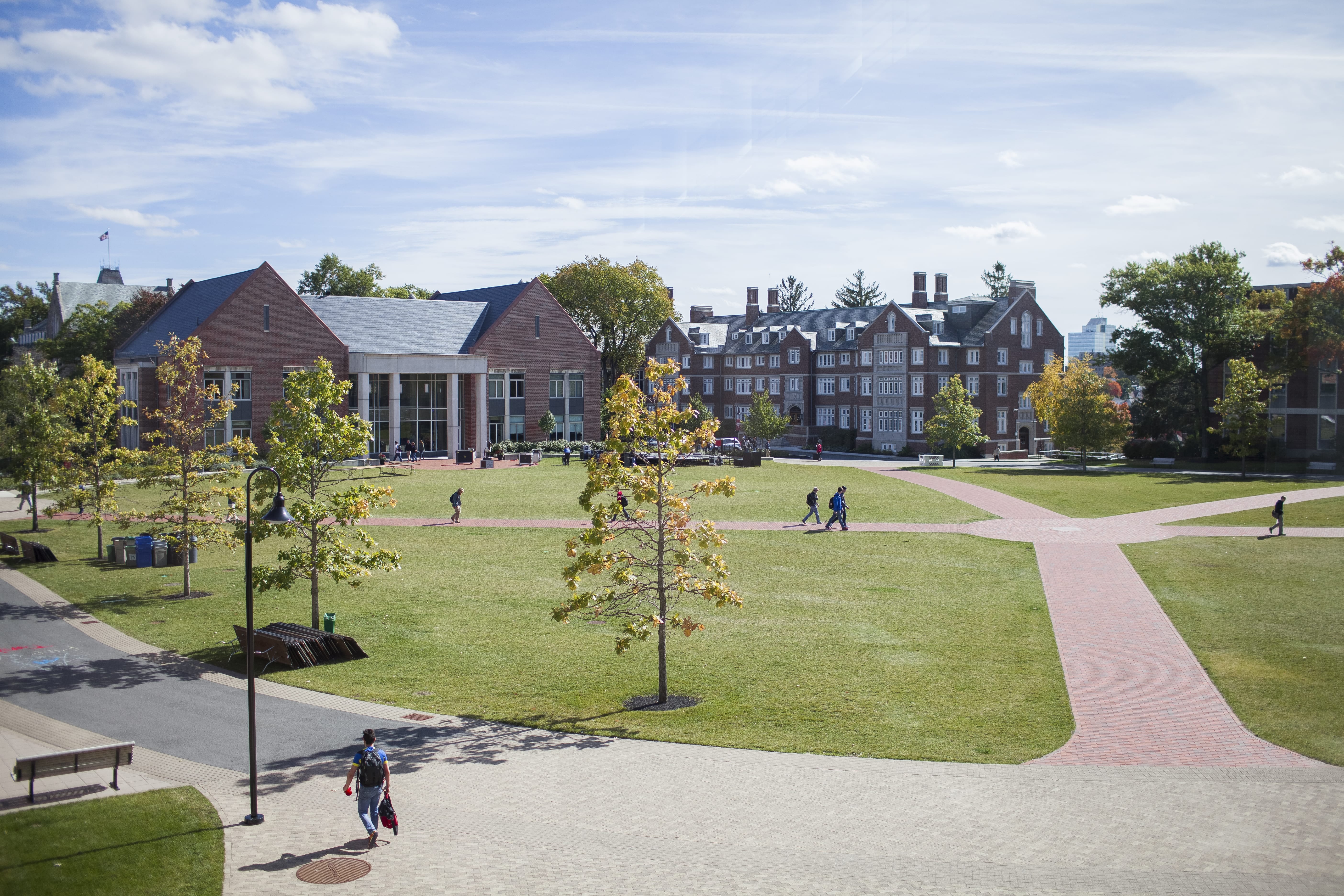 Outside a college campus with brick buildings and plots of green grass in front of them with students walking down the walkways in between the buildings