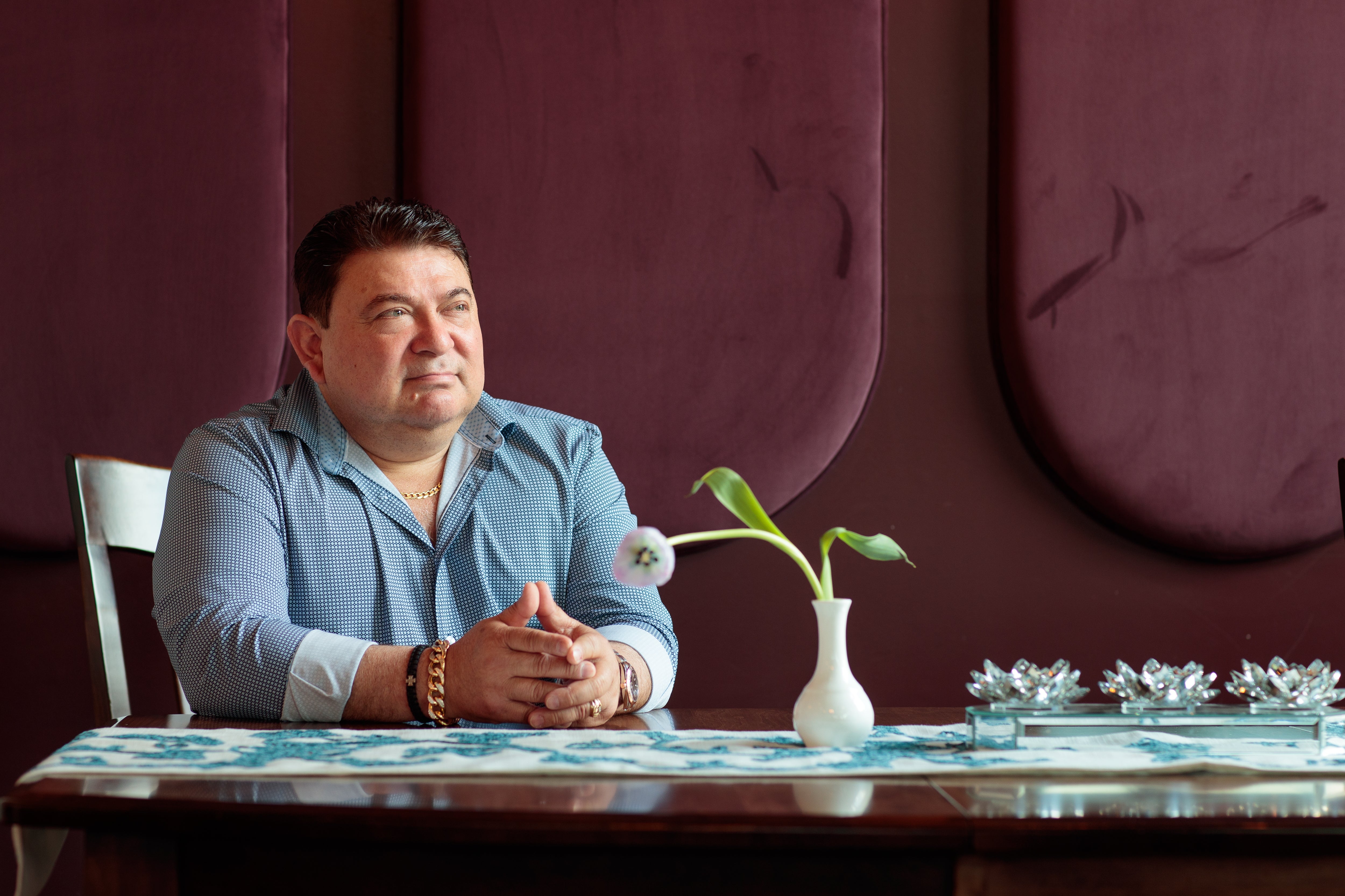 A man with dark hair wears a light blue checkered button down while leaning against a long wooden restaurant table in front of a maroon, textured wall.