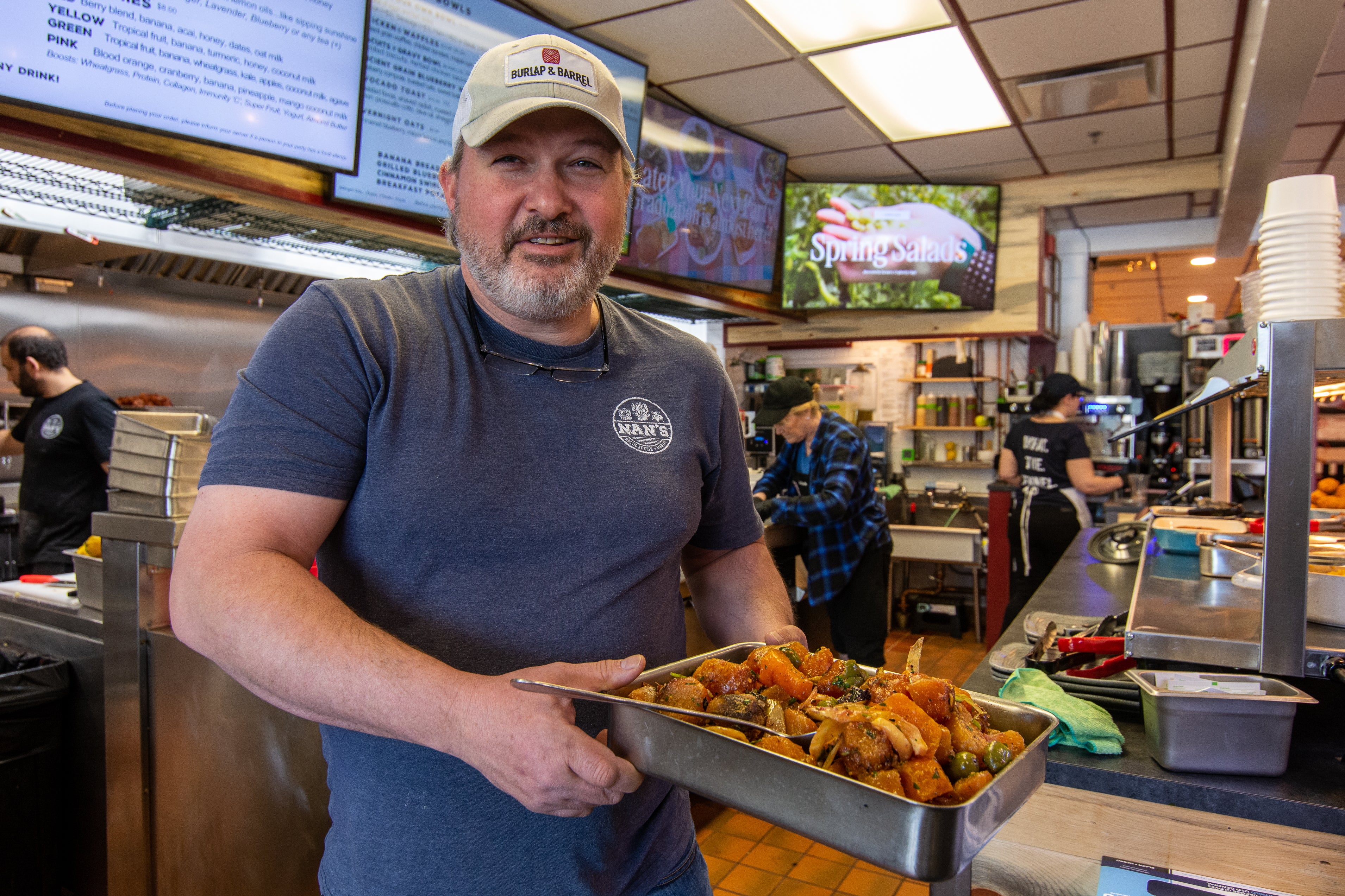 A man wearing a cap and a blue t-shirt holds a tray of cooked vegitables while standing in a restaurant kitchen.